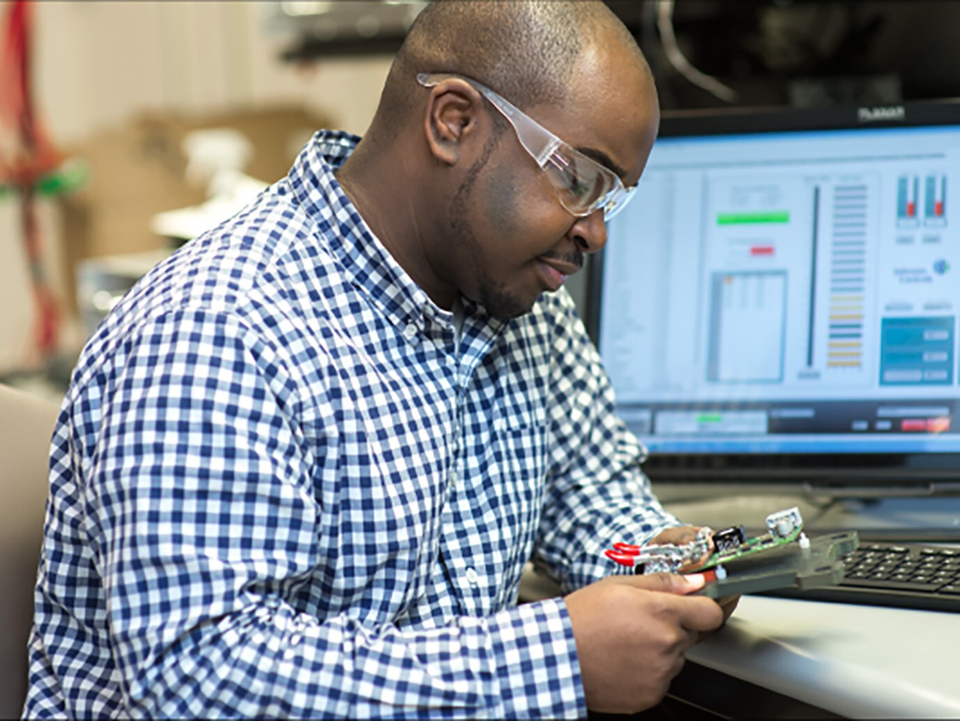 Man wearing safety goggles working with a small device in front of a desktop monitor